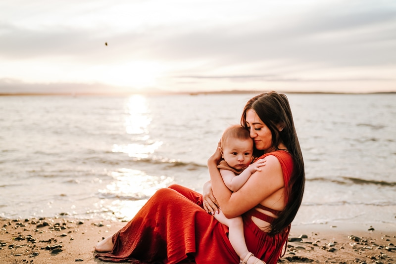 Seattle Motherhood Photography, A woman smiles with grateful joy as she holds onto her baby near the ocean's quiet tide