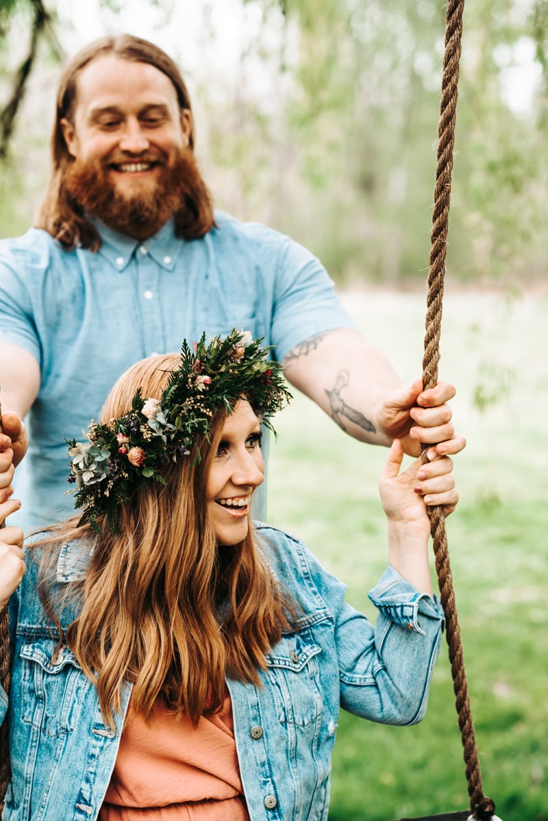 Seattle Couples Photography, a woman sits smiling on a swing with a flower crown, her boyfriend pushes her lovingly