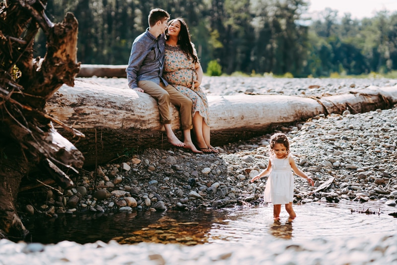 Seattle Maternity Photography, Young husband and wife sit on a long log near a river as their daughter plays
