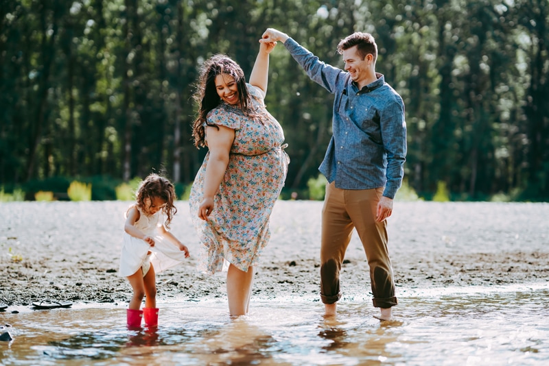 Seattle Maternity Photography, mom, dad, daughter laugh playfully near a river in a forest, dad raises moms hand high