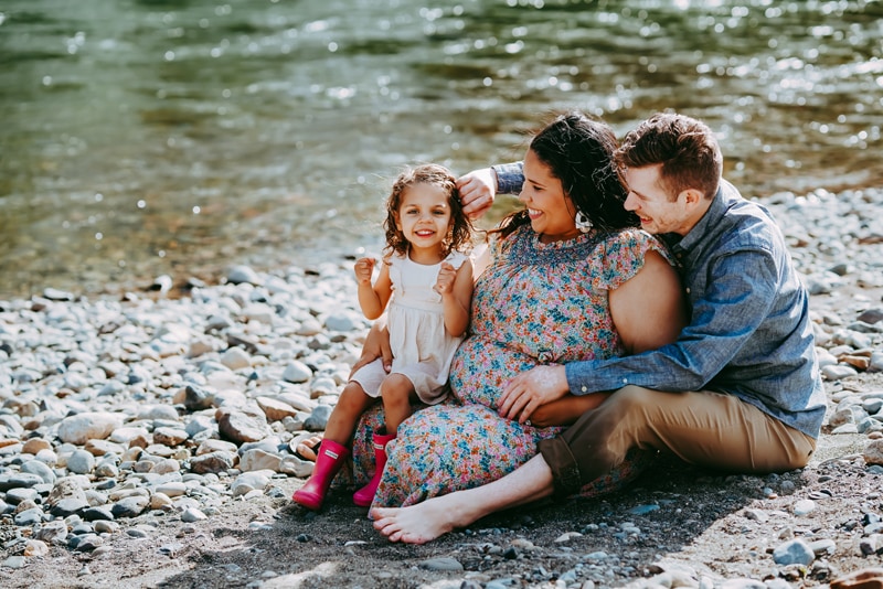 Seattle Maternity Photography, a husband, wife and daughter sit at a river bank, mom is expecting
