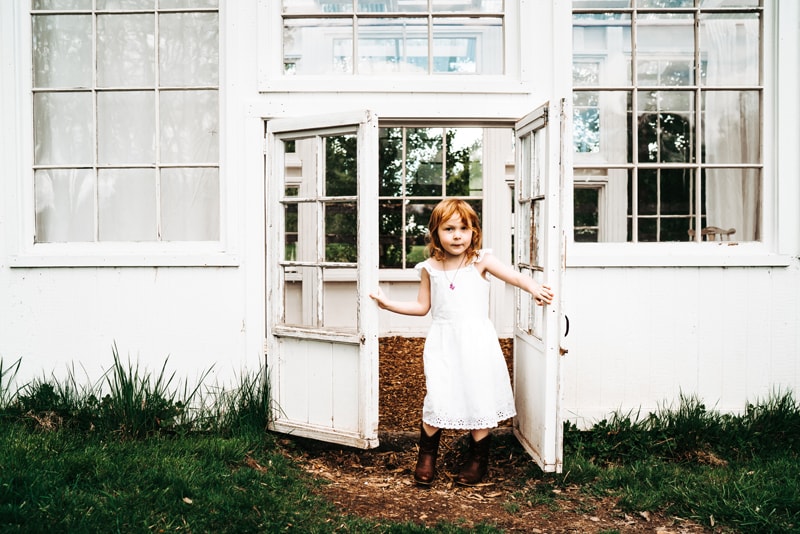 Seattle Motherhood Photography, a young girl opens small white farmhouse doors just her height. She wears boots and a dress.