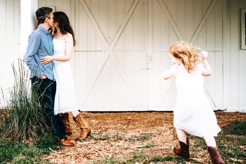 Seattle Motherhood Photography, mom and dad kiss near white barn doors outside, their daughter runs toward them