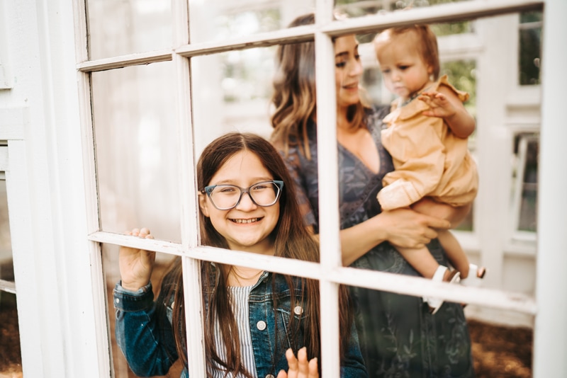 Seattle Motherhood Photography, through a window frame a young girl smiles, her mother holds baby sister smiling close by