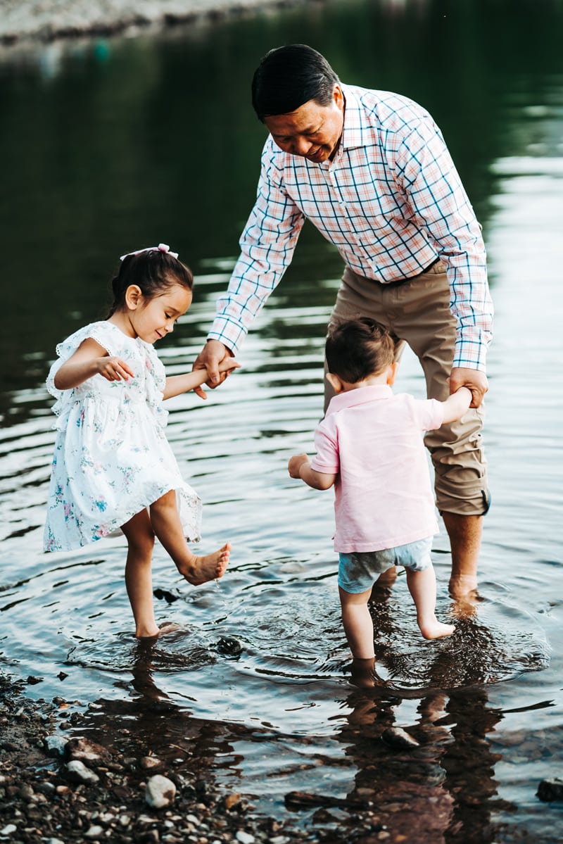 Seattle Grandparent Legacy Photography, a grandfather holds his grandchildren's hands on a lakeshore, toes in the water