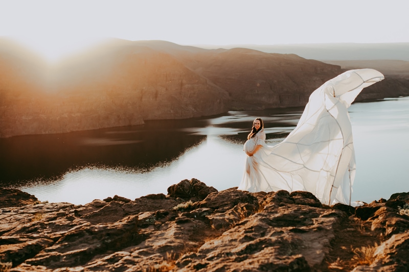 Seattle Maternity Photography, pregnant woman in a white dress stands outdoors in nature, the train of her dress doubles her height as it lifts into the air by the wind