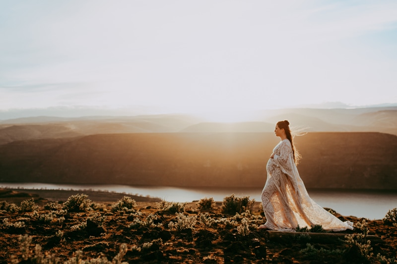 Seattle Maternity Photography, a pregnant mother in a lacy white dress walks along a hillside near the river