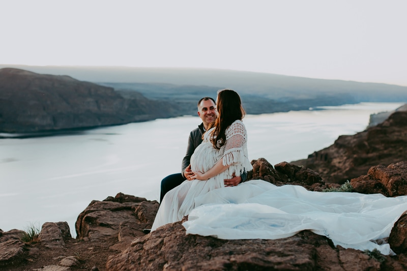 Seattle Maternity Photography, a very pregnant woman in a long flowing white dress sits with her husband near a large river