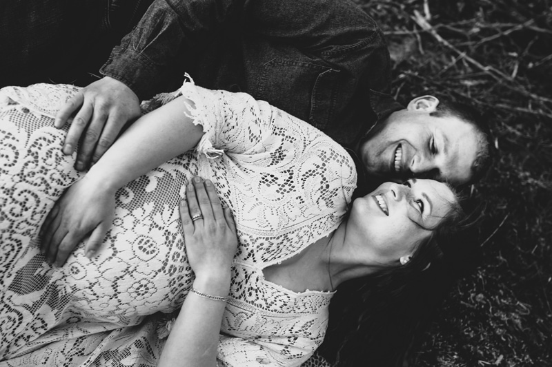 Seattle Maternity Photography, man holds his pregnant wife as she looks back at him, they lay in the grass