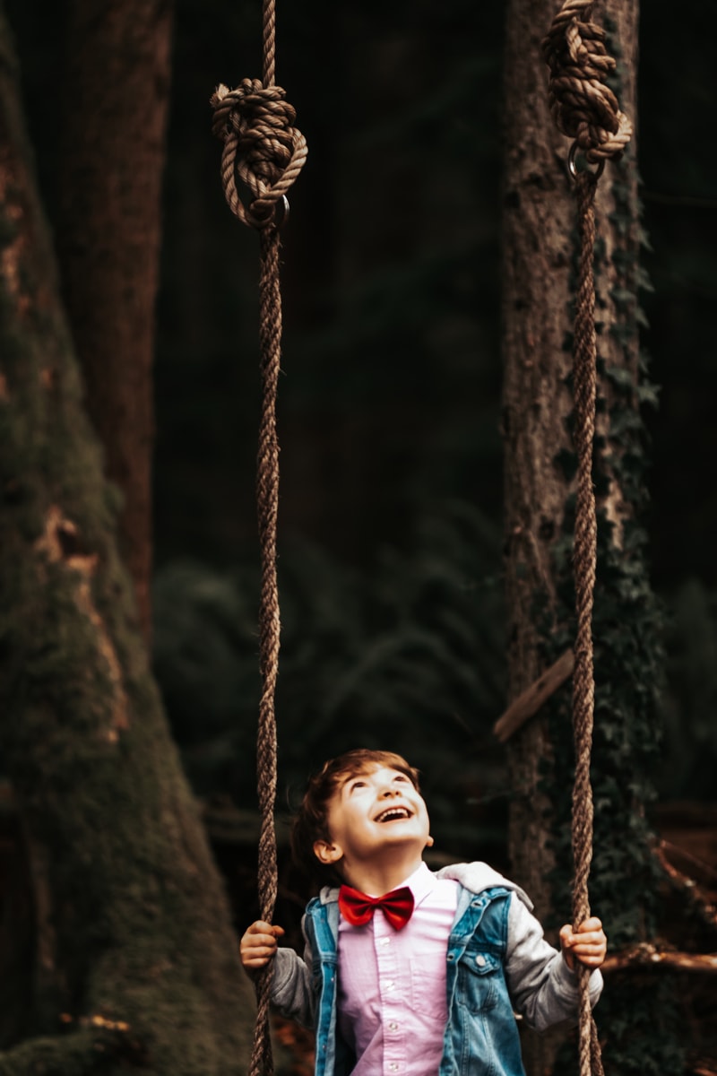 Seattle Motherhood Photography, a young boy smiles, his bow tie on as he sits on a tree swing in the forest