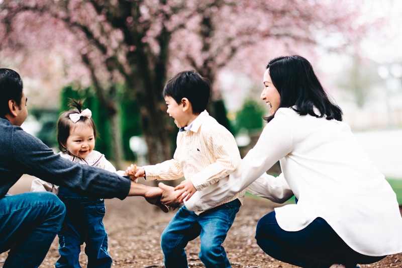 Seattle Motherhood Photography, a young family of four all hold hands and smile underneath a cherry blossom tree