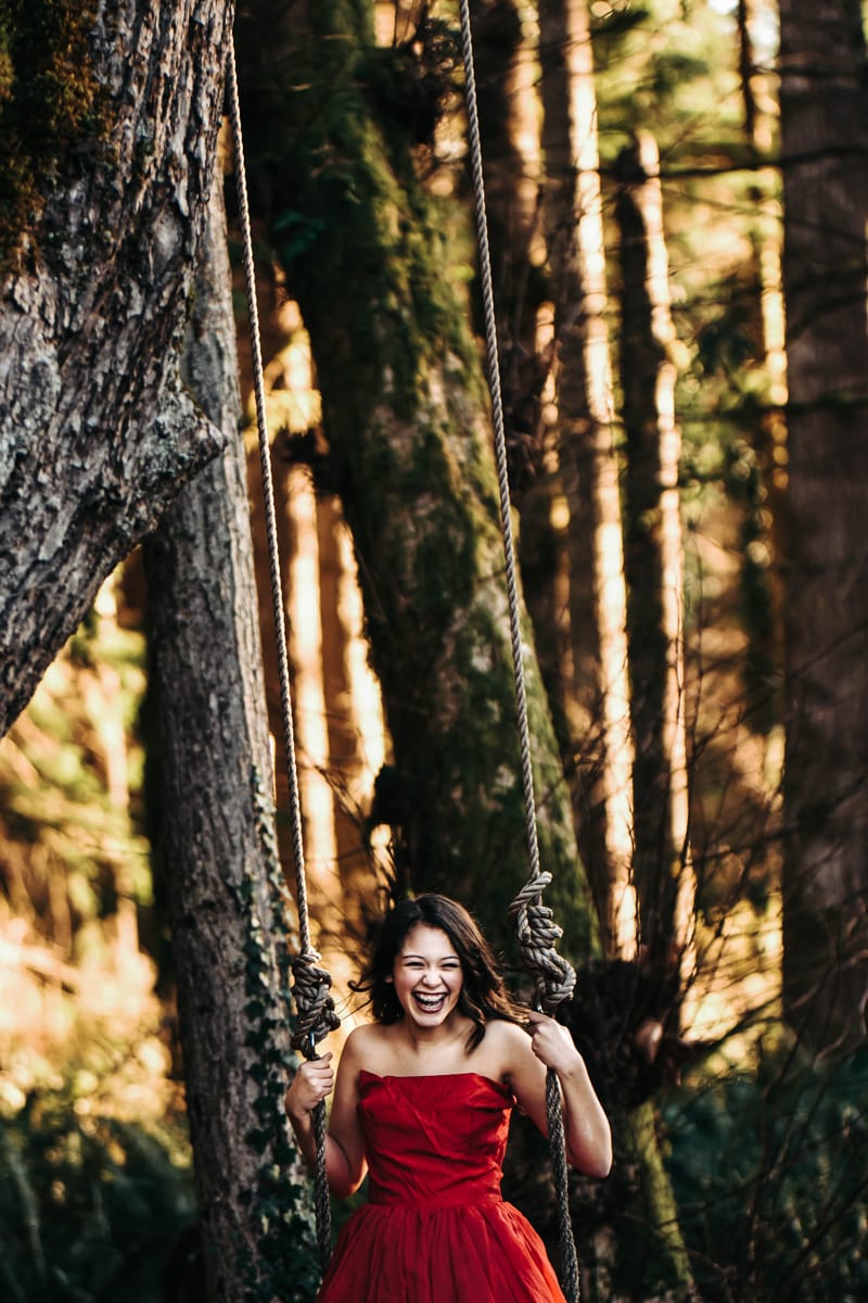 Seattle Senior Photography, a girl in a red dress laughs and smiles excitedly as she enjoys a swing in the forest