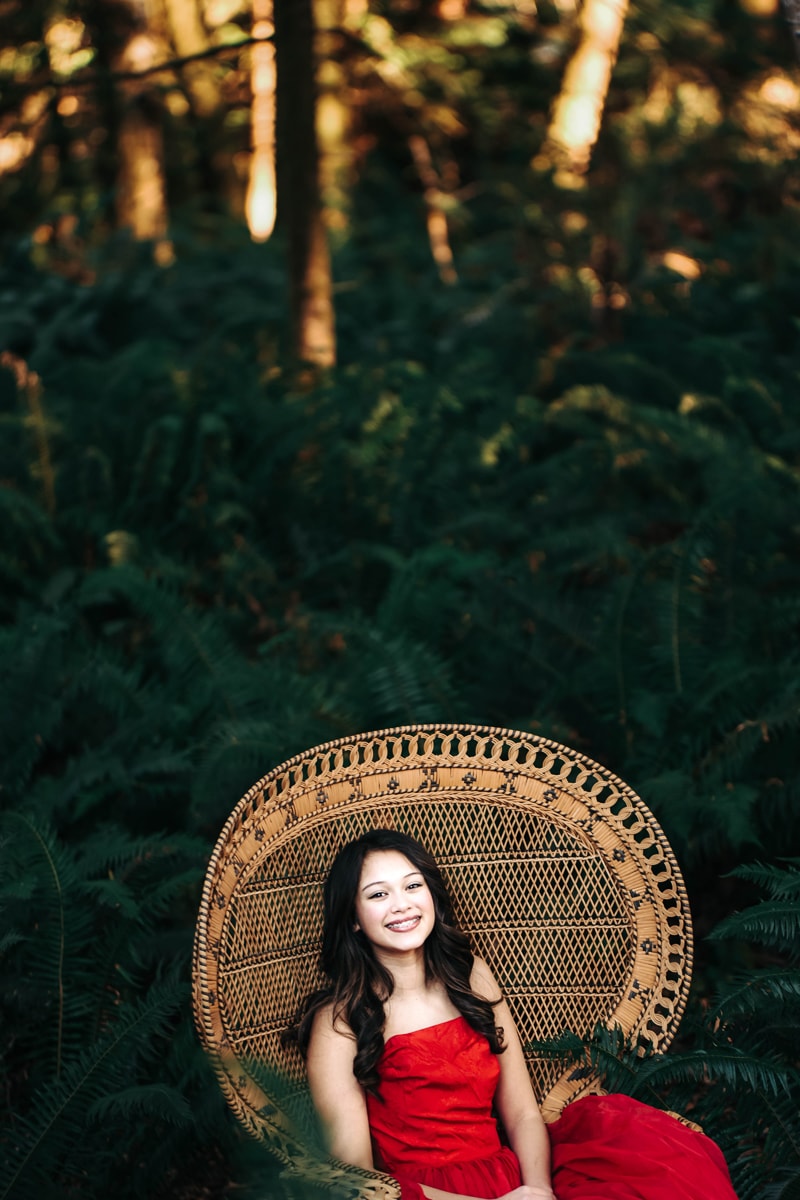Seattle Senior Photography, a high school girl sits in a wicker chair before a forest, she smiles