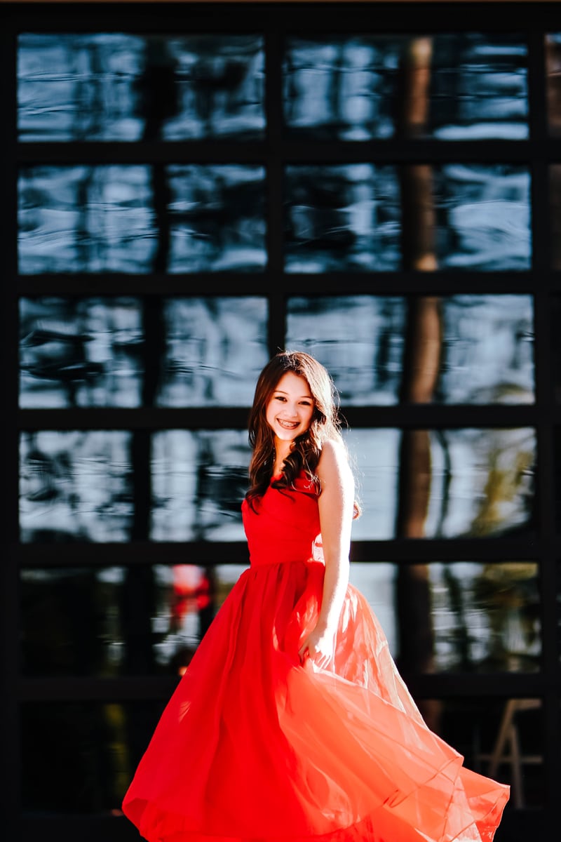 Seattle Senior Photography, a high school woman in a flowing red dress stands outside near reflective windows, the sun shines on her back